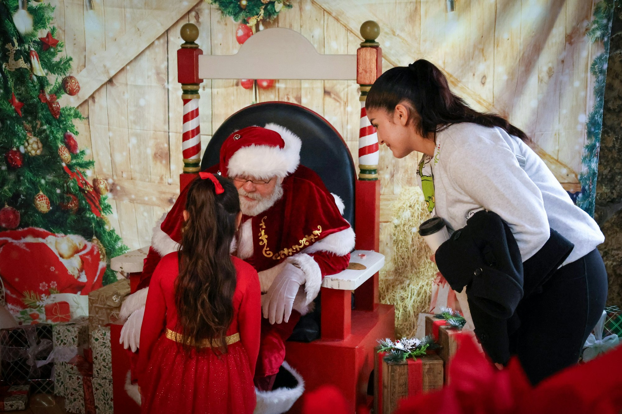 A young girl in a red dress talks to Santa Claus while another woman watches, surrounded by festive decorations and gifts.