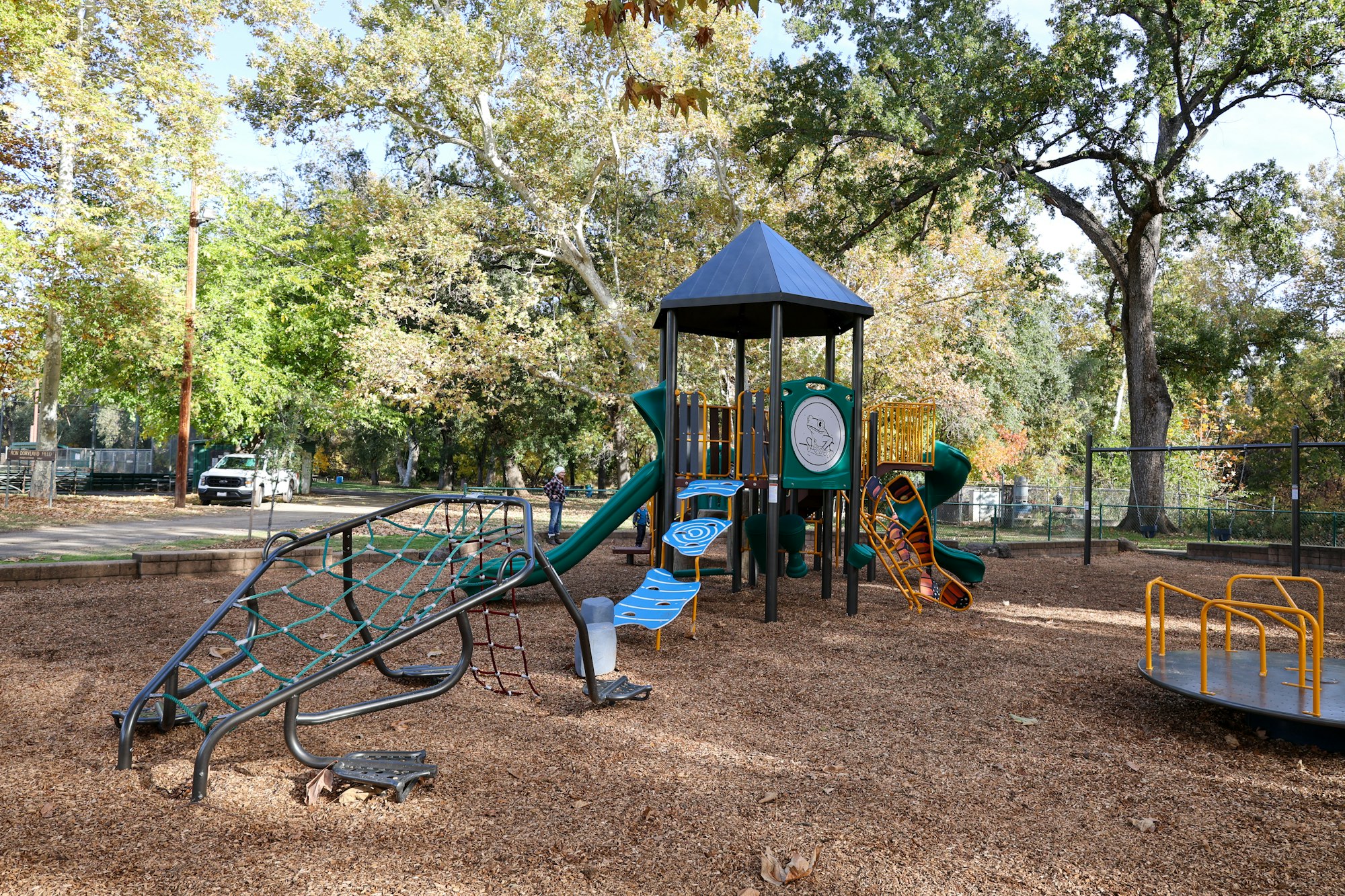 A playground with slides, a climbing net, and a merry-go-round, surrounded by trees.