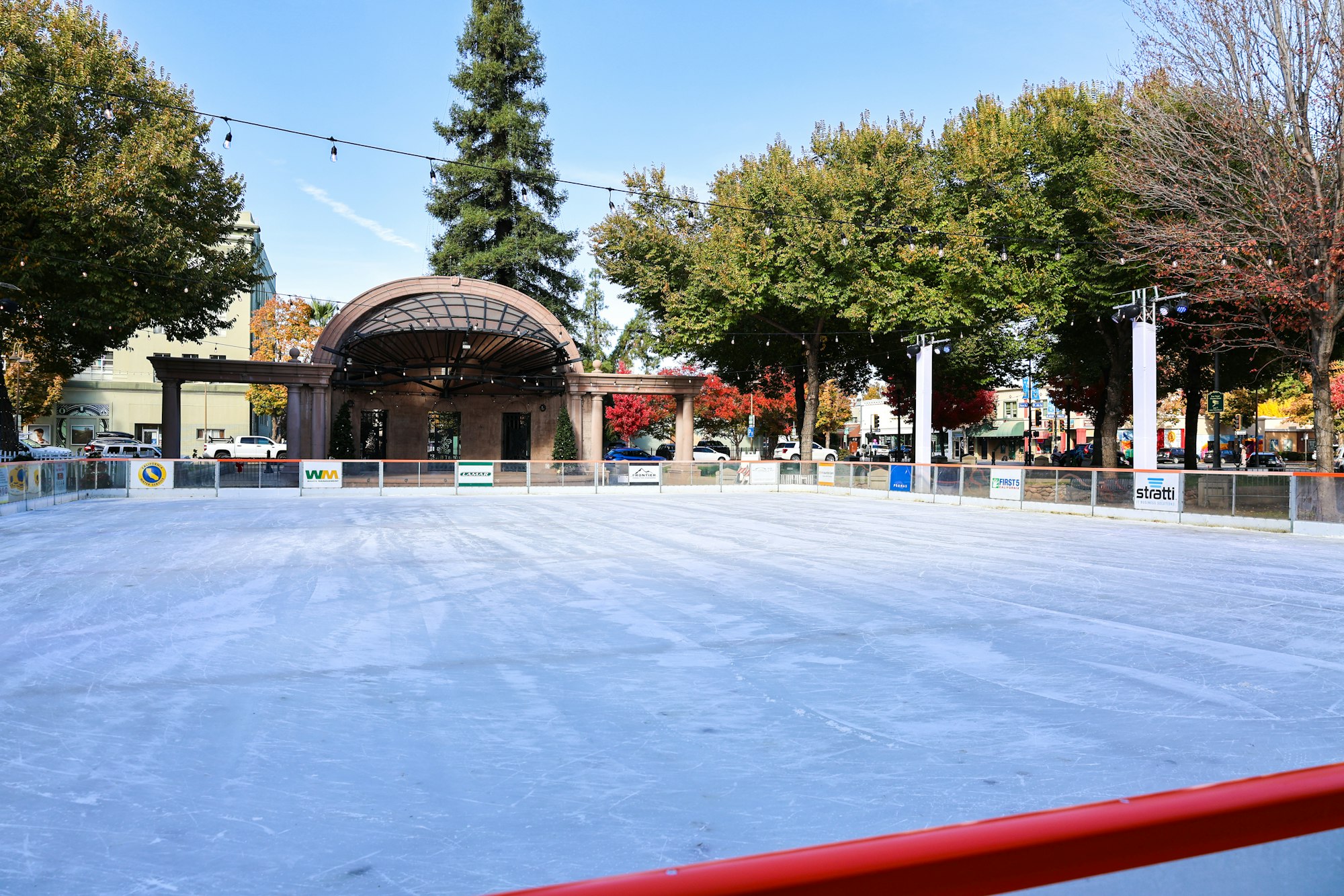 An empty outdoor ice skating rink with trees and a gazebo-like structure in a park setting.