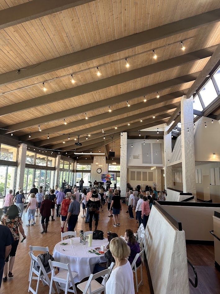 A lively gathering in a spacious venue with people dancing, tables set, and large windows letting in natural light.