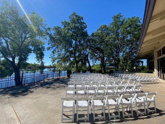 Outdoor event setup with rows of white chairs on a patio near trees and a river, under a clear blue sky.