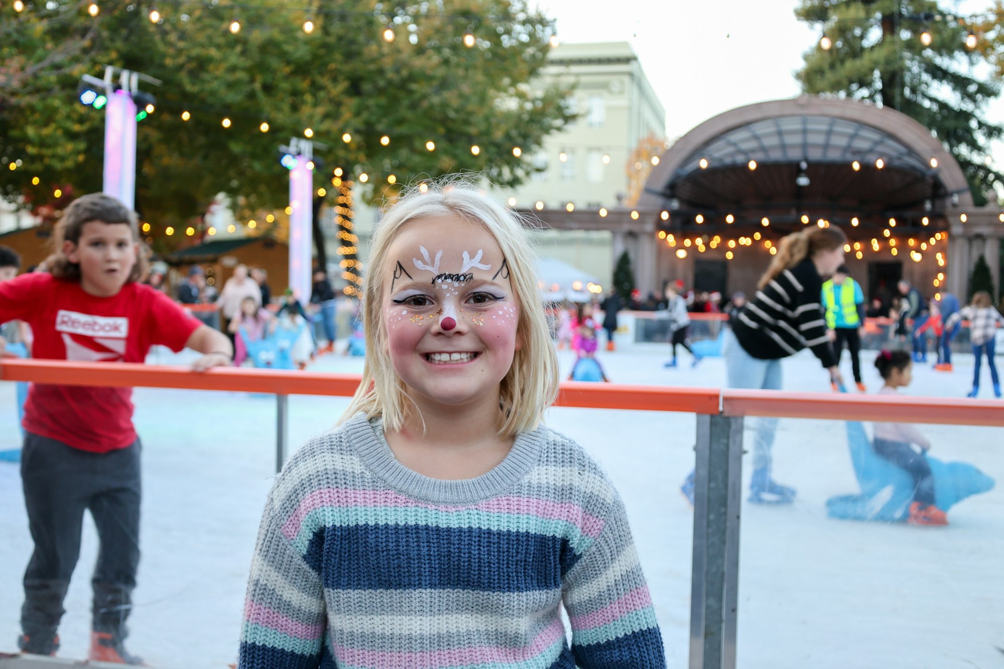 A happy child with face paint in front of an ice-skating rink with lights and skaters.