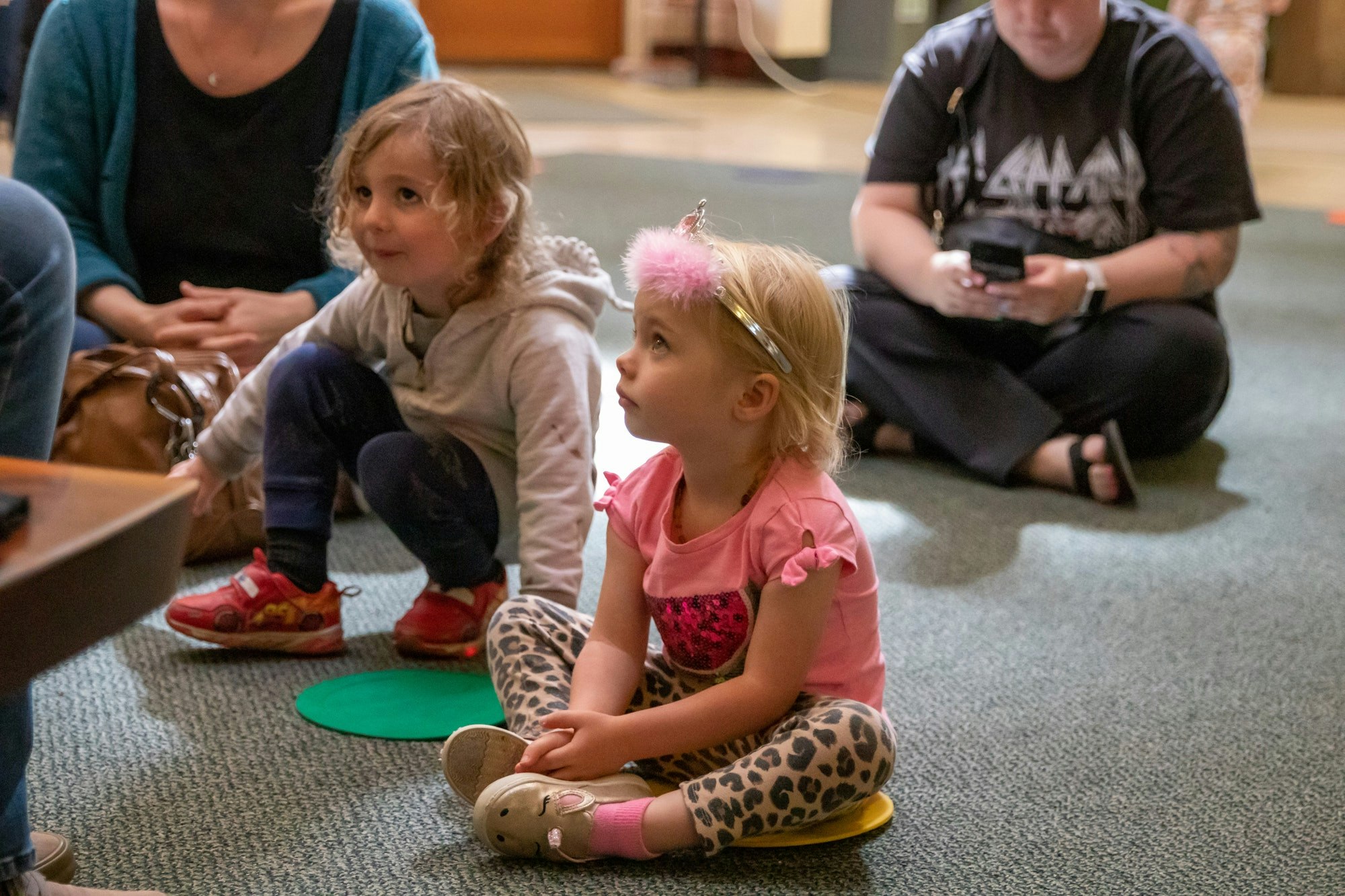 Two young children sitting on the floor, engaged and listening, with adults in the background. The setting appears cozy and informal.