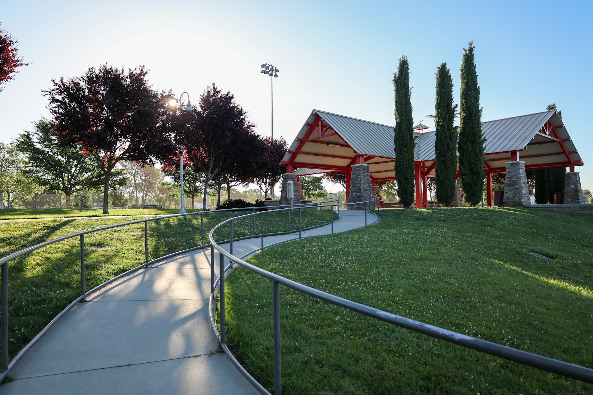 A park with a winding pathway, green grass, trees, and a red-roofed pavilion under a clear sky.