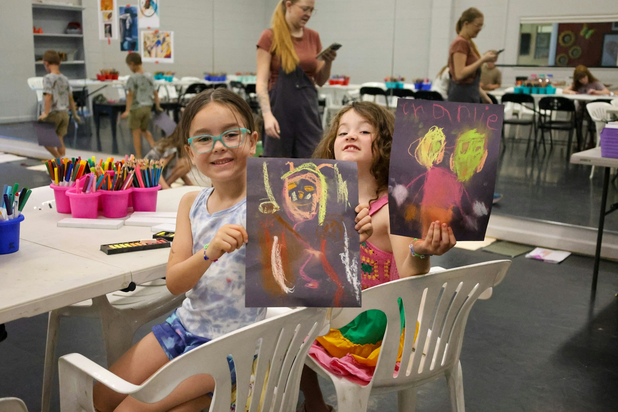 Two smiling girls hold up colorful artwork in a creative classroom filled with art supplies and other children.