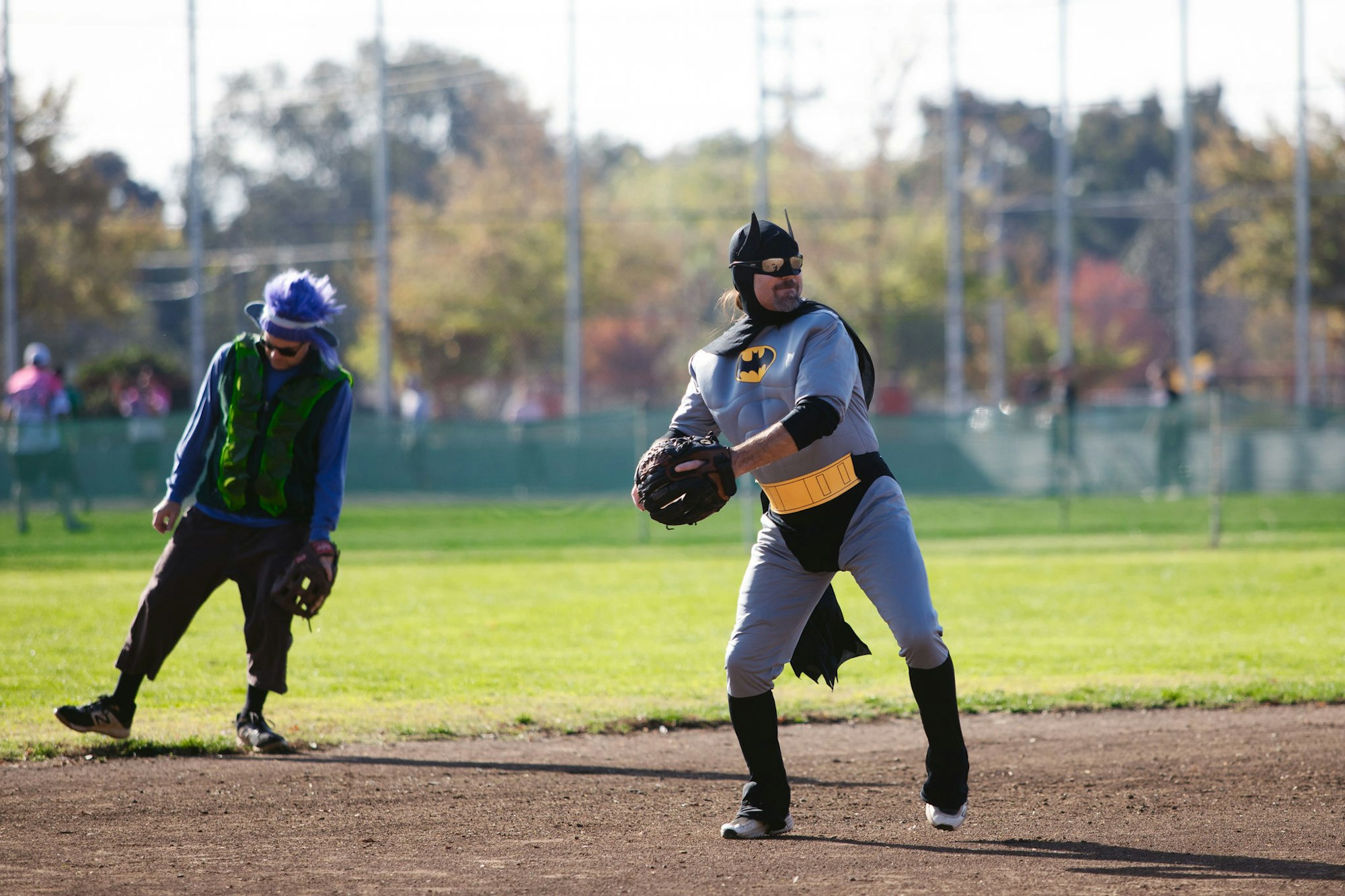 Person dressed as Batman playing baseball with another in a colorful wig and sunglasses.