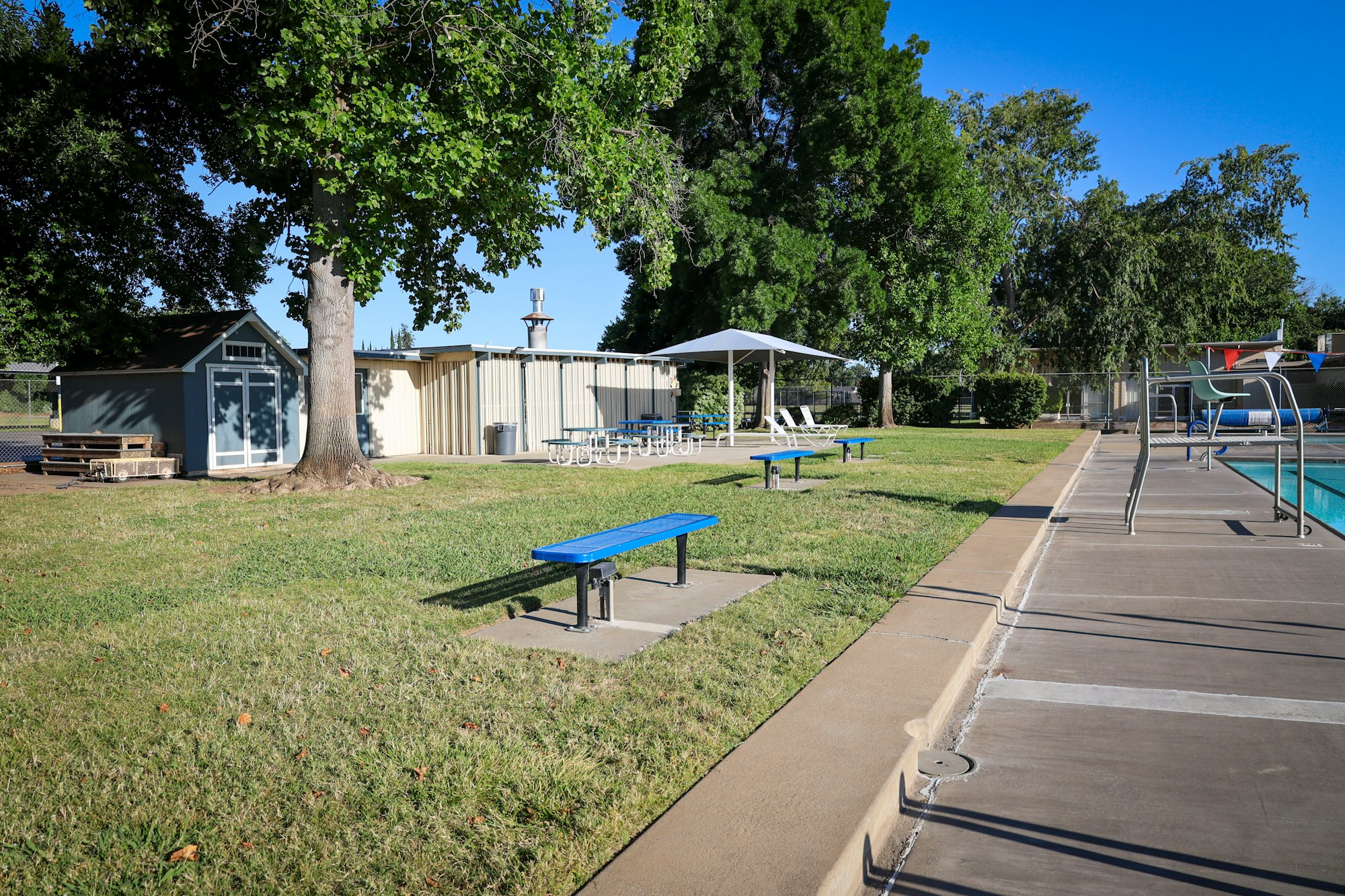 Poolside area with benches, a pool, a building, tables, chairs, and trees on grass.