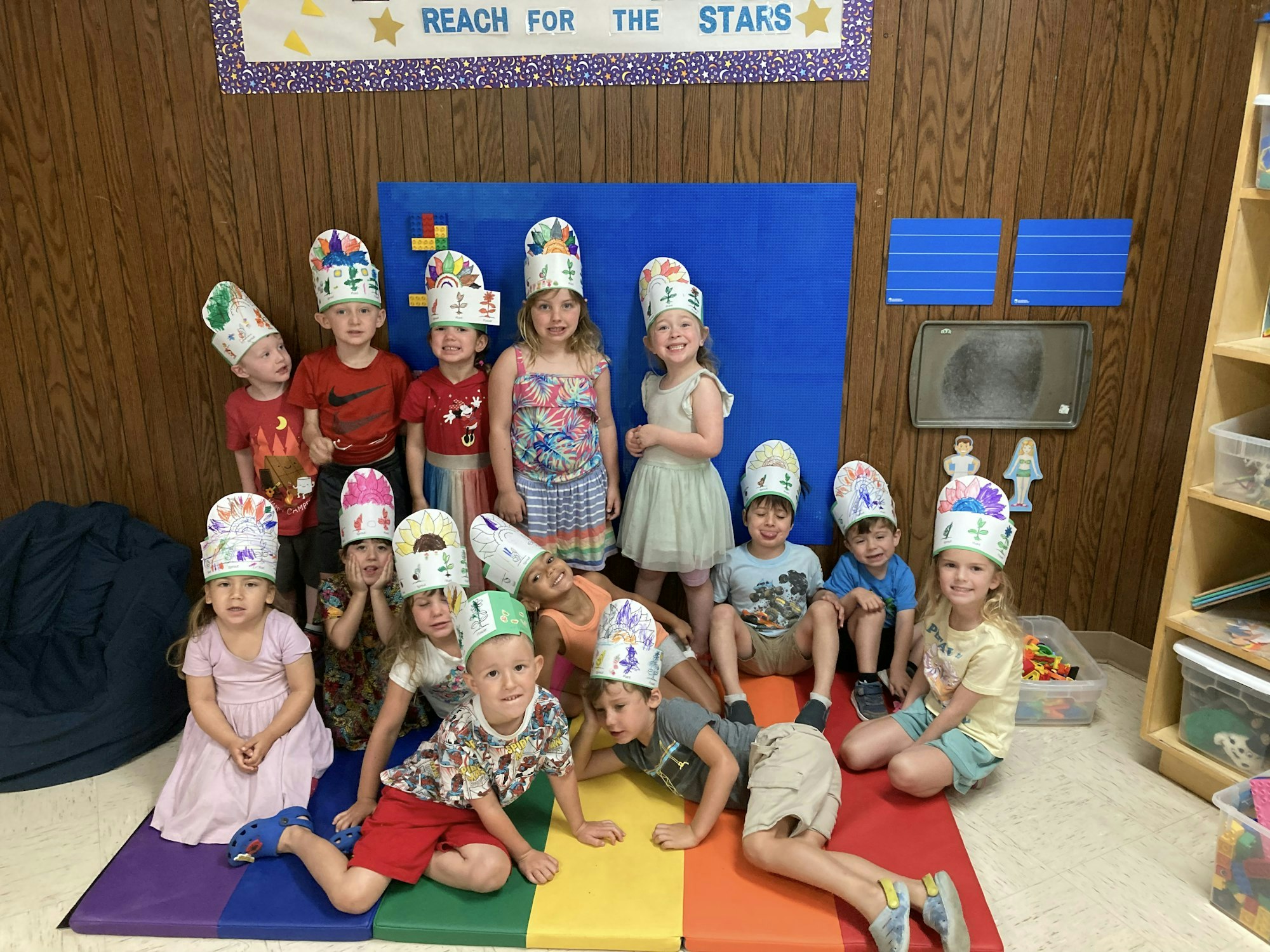 A group of children wearing colorful, decorated hats sit together on a mat in a classroom, smiling and posing for the photo.
