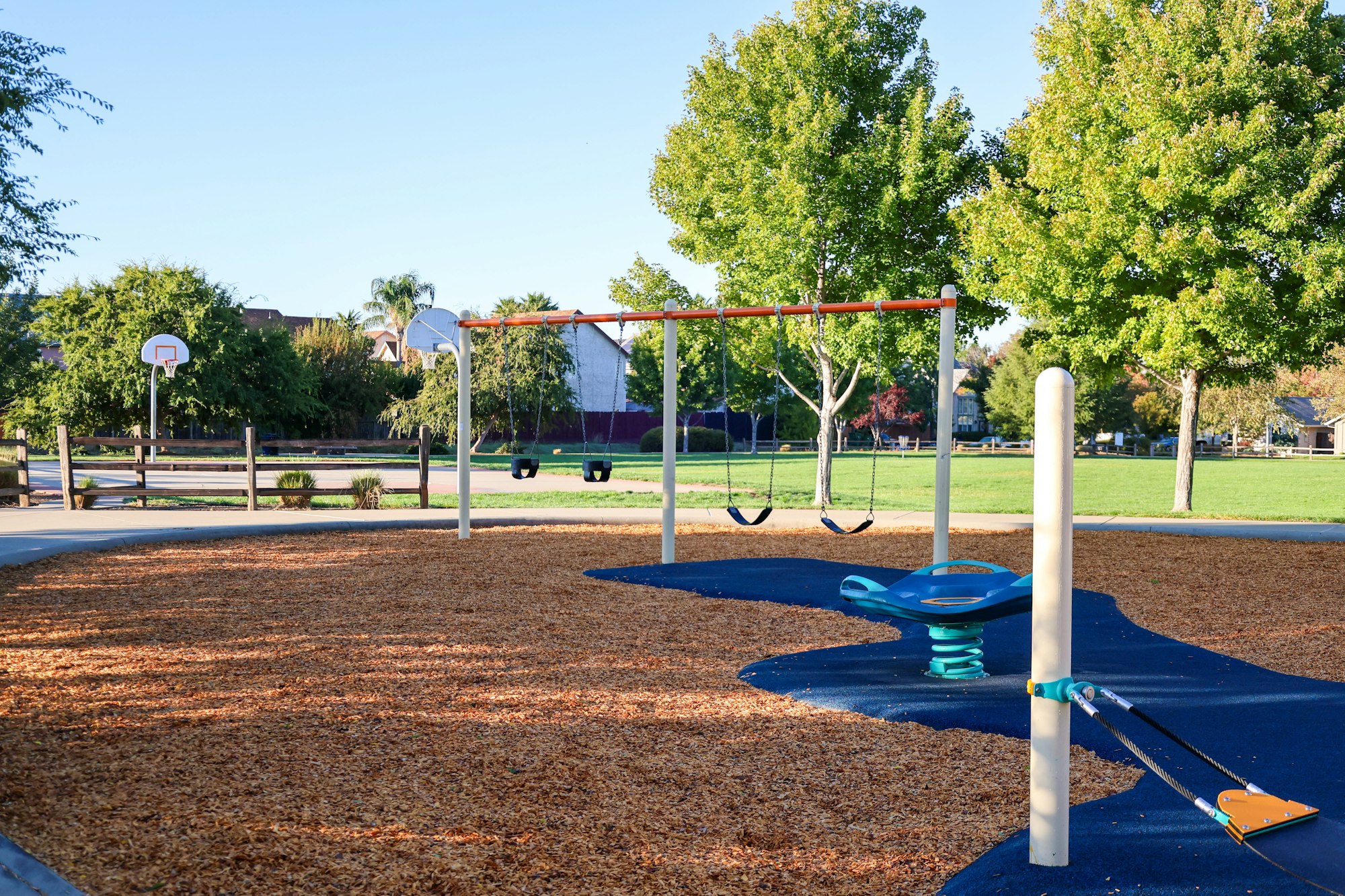 A playground with swings, a seesaw, woodchips, and nearby basketball court, surrounded by trees and grass.