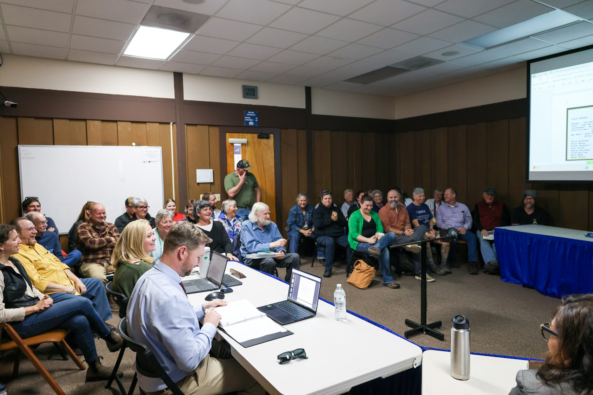 A group of people in a meeting room, some seated, with a presentation visible on a screen.