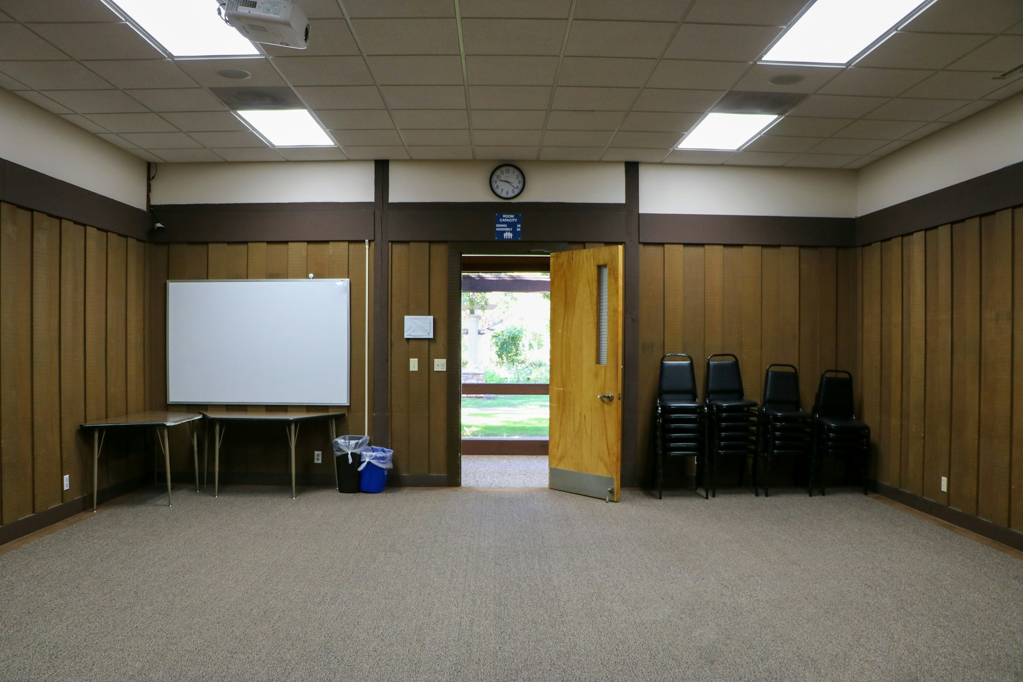 An empty meeting room with a whiteboard, chairs stacked to the side, and an open door showing outdoor greenery.