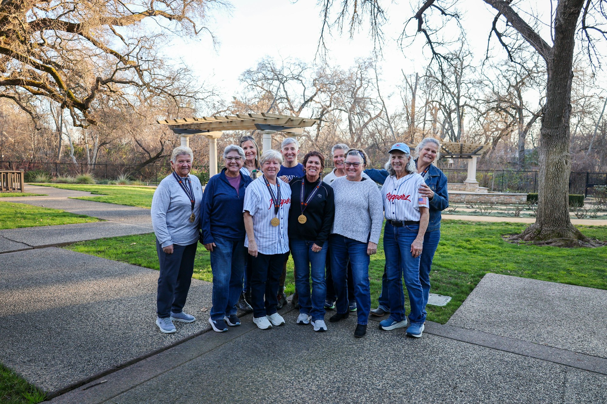 A group of people posing outdoors, some in baseball jerseys, with medals around their necks, in a park-like setting.