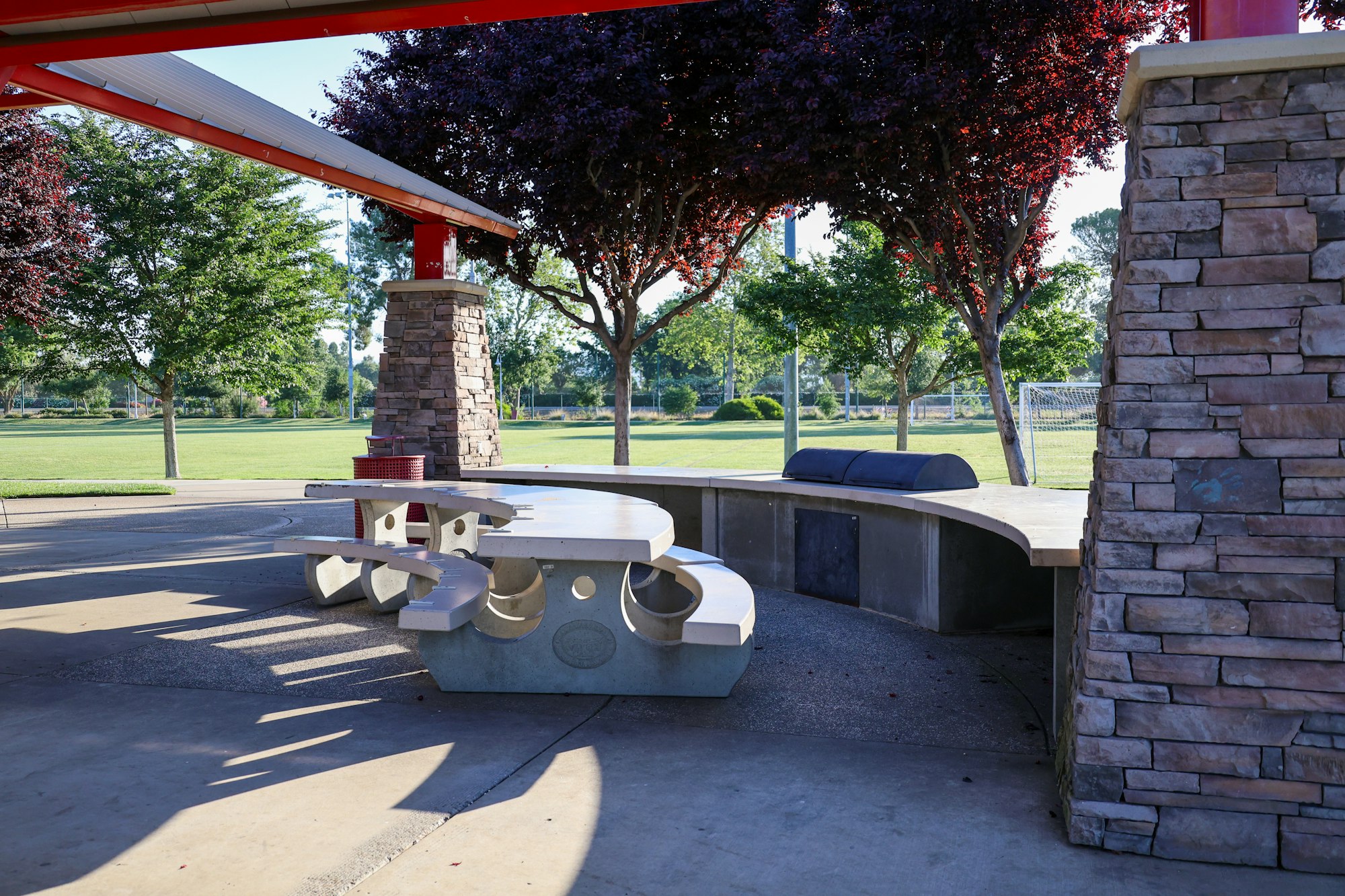 Picnic area with concrete tables, a BBQ grill, trees, and a glimpse of a soccer goal in a park.