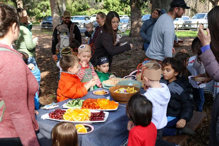A group of children and adults enjoying a picnic outdoors, with a variety of snacks and fruits on a table.