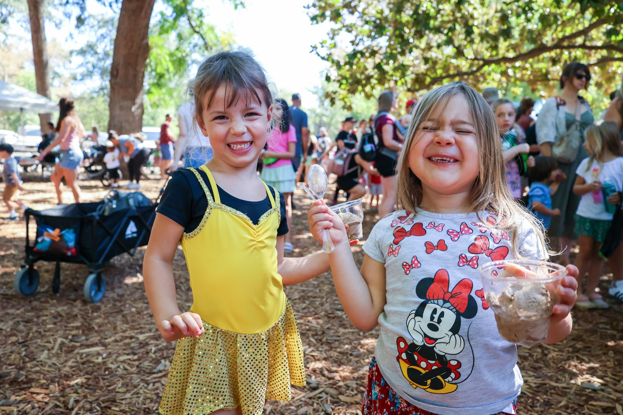 Two kids smiling, one in a yellow dress, the other holding ice cream, outdoors with people and trees in the background.