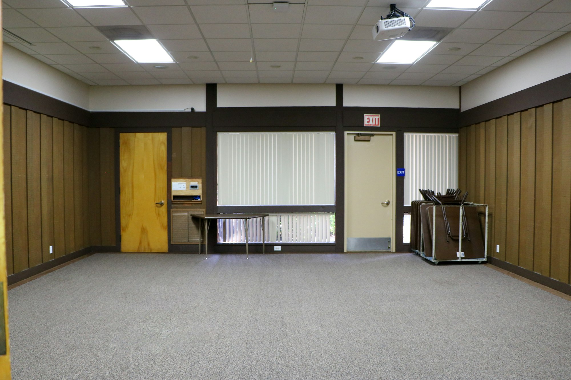 An empty room with a wood door, windows with blinds, projector, carpet flooring, and a cart with folded chairs.