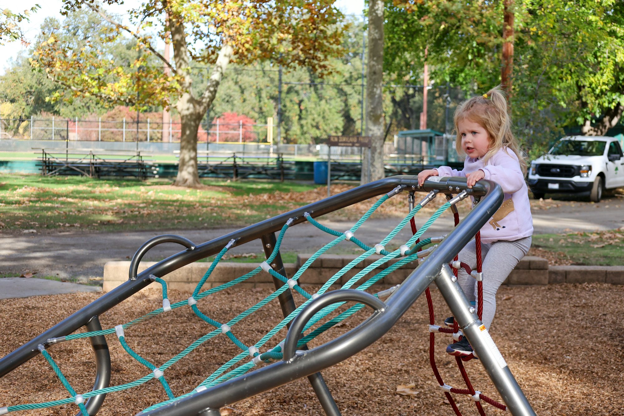 A child playing on a rope climbing structure at a playground, with trees and park benches in the background.