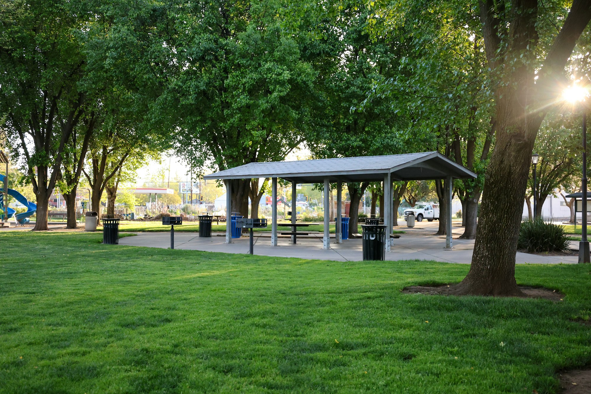 A park with trees, a picnic shelter, benches, and a playground in the background, illuminated by a setting sun.