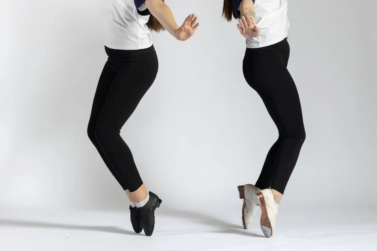 Two dancers performing a tap dance pose, wearing black leggings and contrasting tap shoes, set against a neutral background.