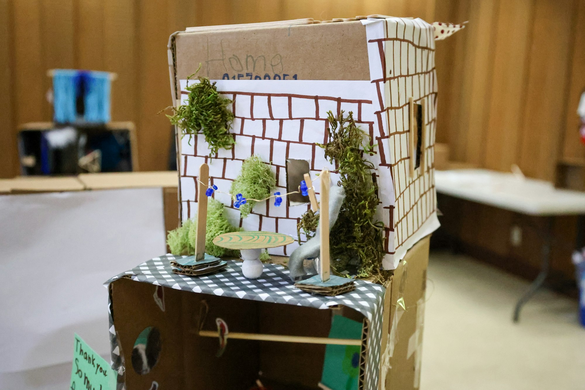 A small, handcrafted model house made from cardboard and moss, with a mini table and string decorations.