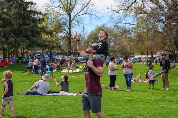 A dad carries his son on his shoulders, both looking up in a lively park filled with families and children flying kites.