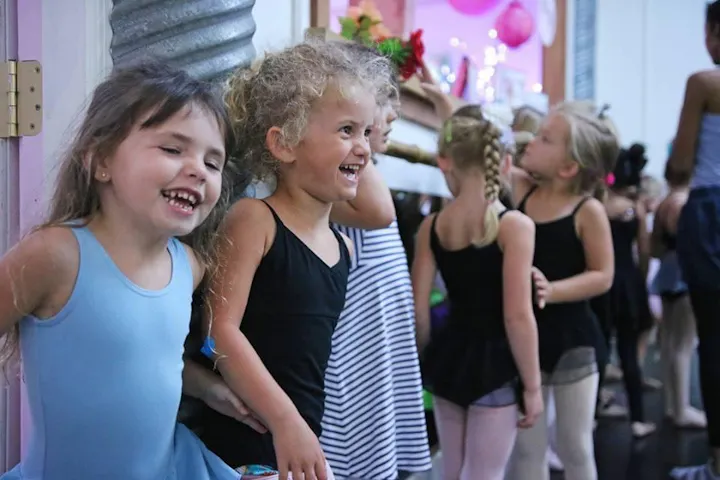 A joyful group of young girls in dance attire smiles and interacts, capturing a fun moment in a dance studio setting.