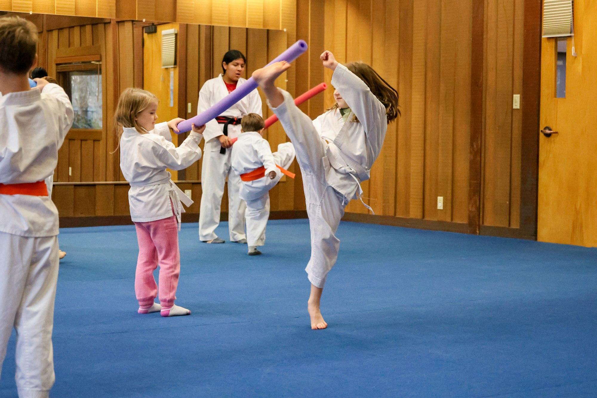 Children in a martial arts class practicing with foam swords, instructor watching.