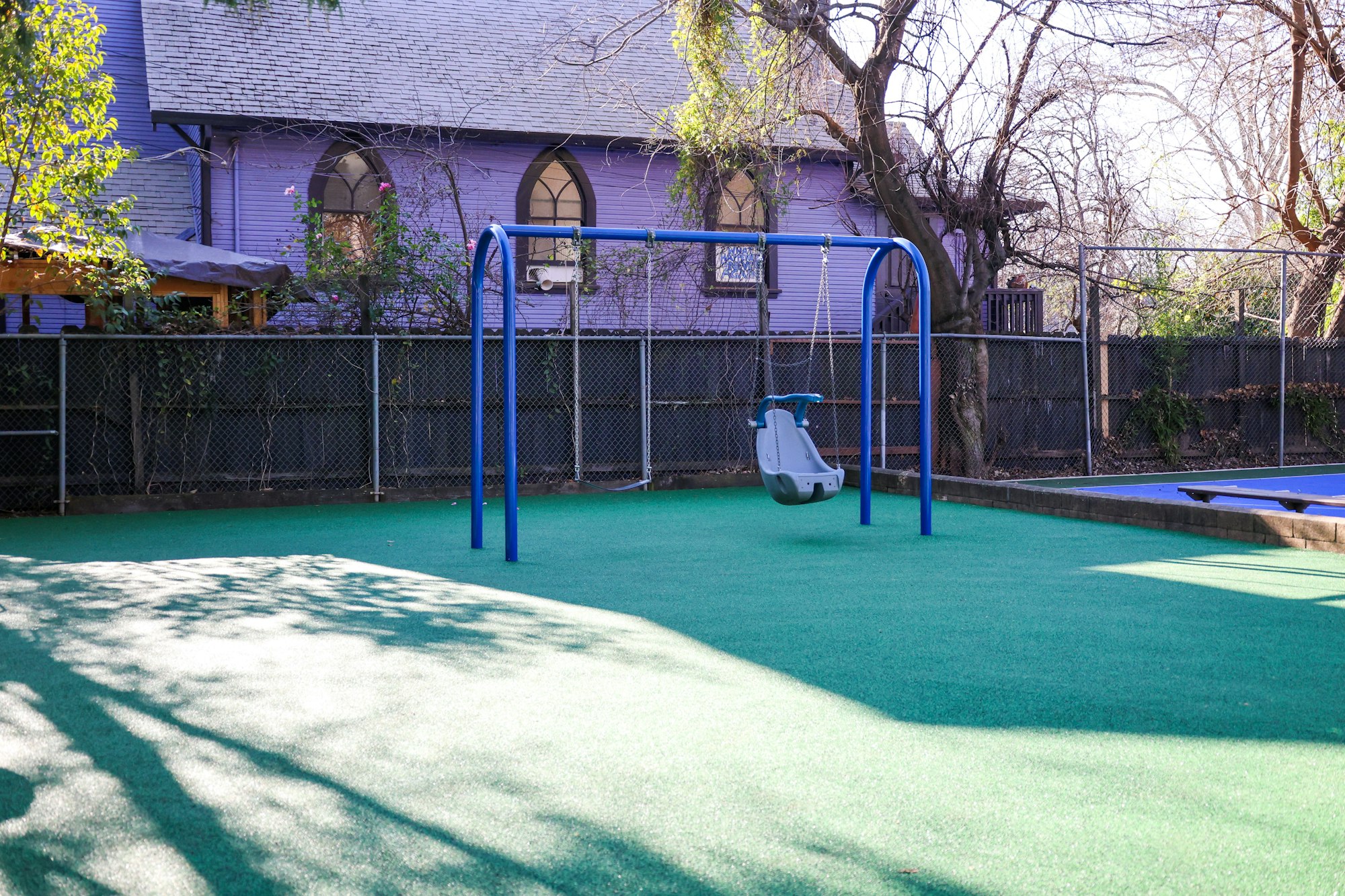 Playground with a single swing, green turf, fence, and purple building in the background.