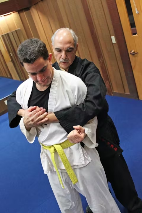 A martial arts practice scene with one instructor demonstrating a technique on a student wearing a yellow belt.
