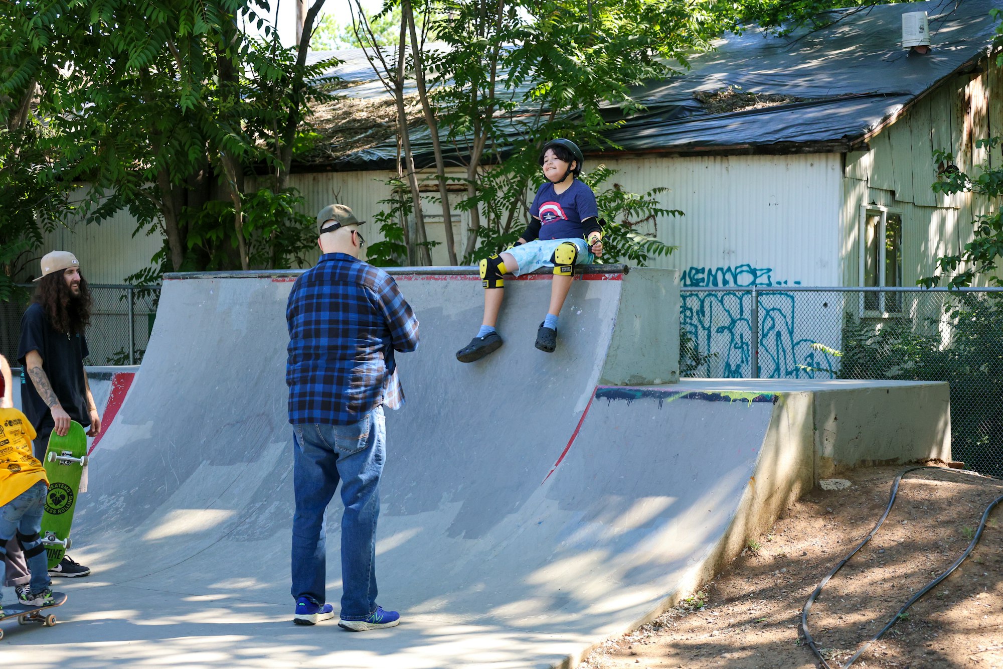 A child sits atop a skate ramp while adults with skateboards stand by, amidst an urban backdrop with graffiti.