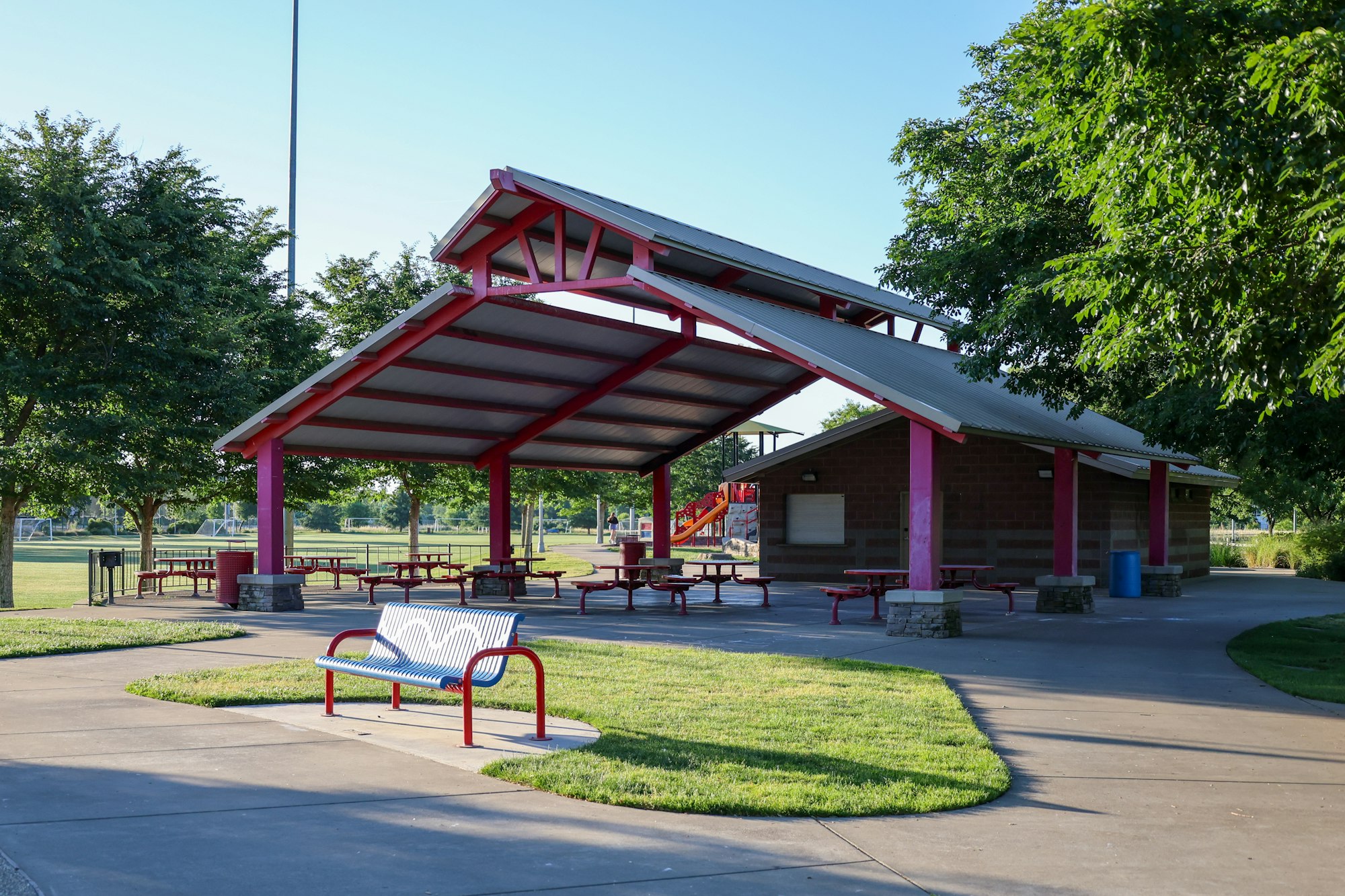 A park pavilion with picnic tables, a playground slide in the background, a bench, and green trees under a clear sky.