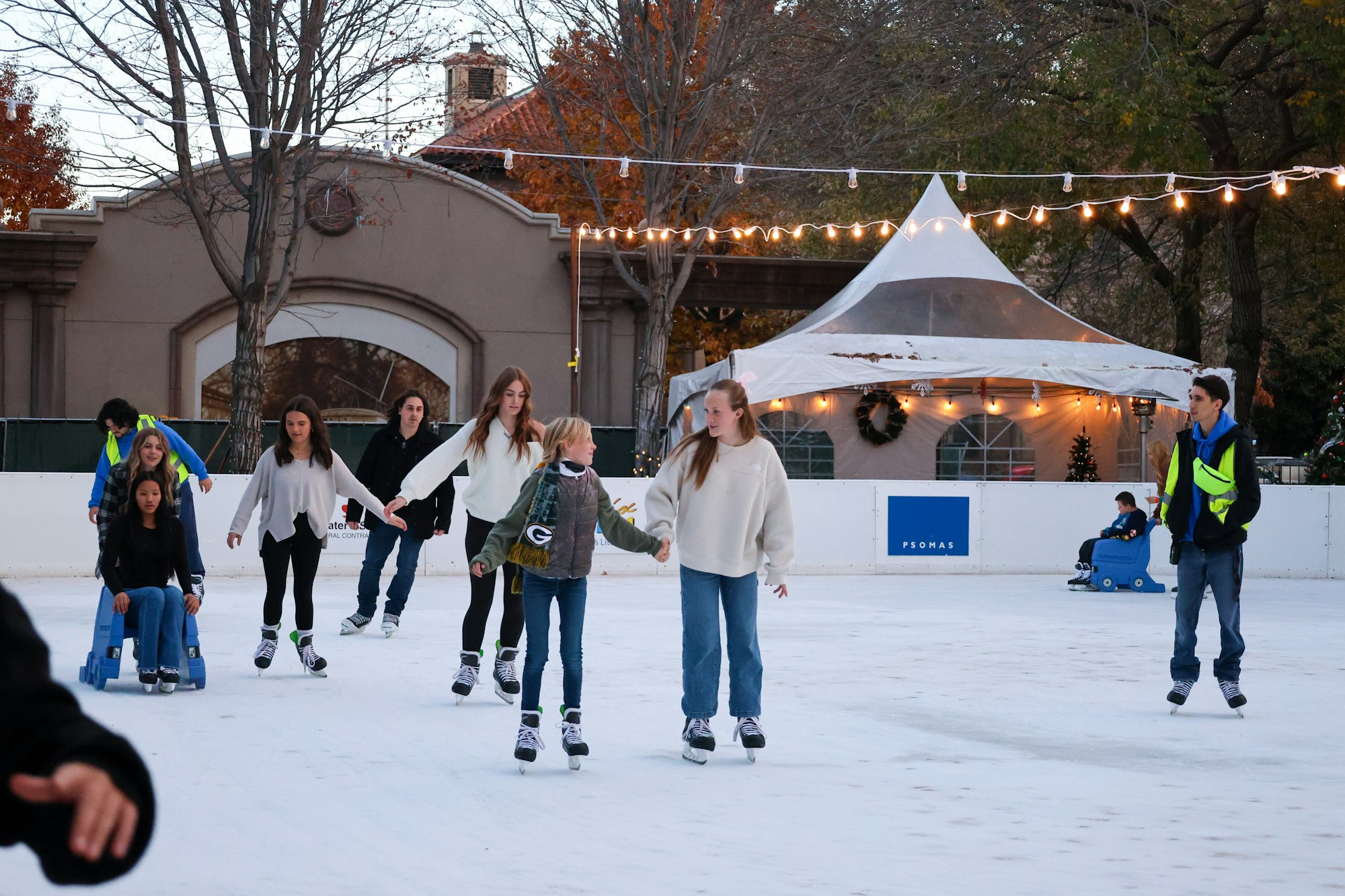People ice skating in an outdoor rink, with festive lights and a tent in the background.
