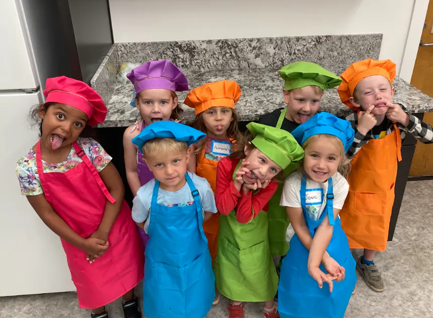 A group of children wearing colorful chef hats and aprons poses playfully in a kitchen.