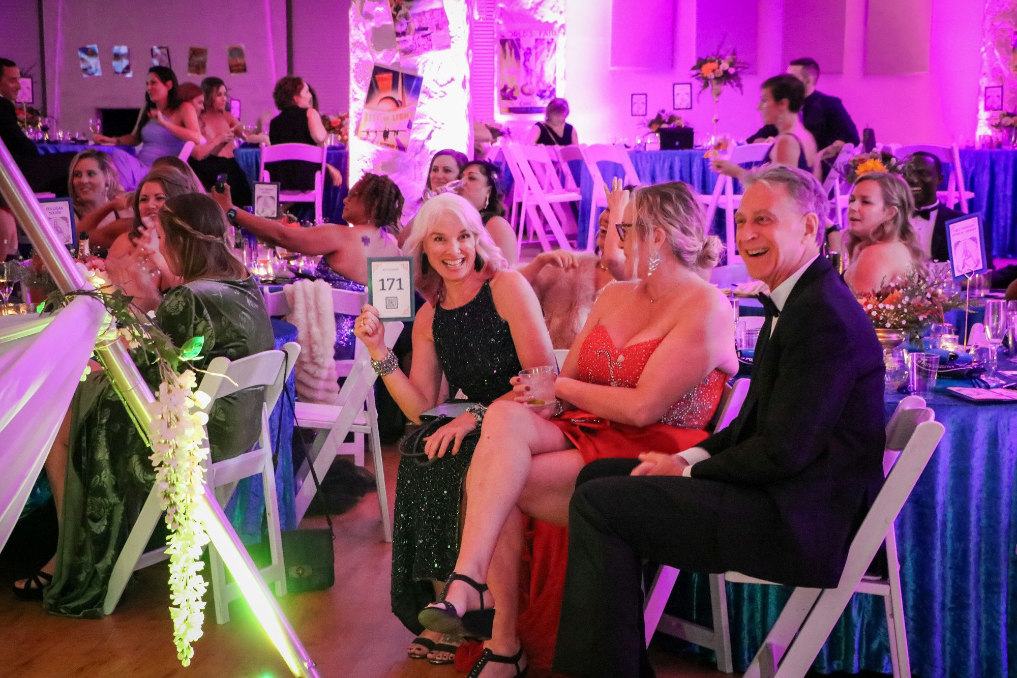 A lively event with people in formal attire, a woman holding a "171" card, and colorful decor in a festive setting.