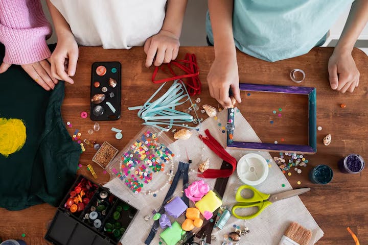A messy craft table with hands visible, showcasing various art supplies, beads, colorful materials, and a decorated T-shirt.