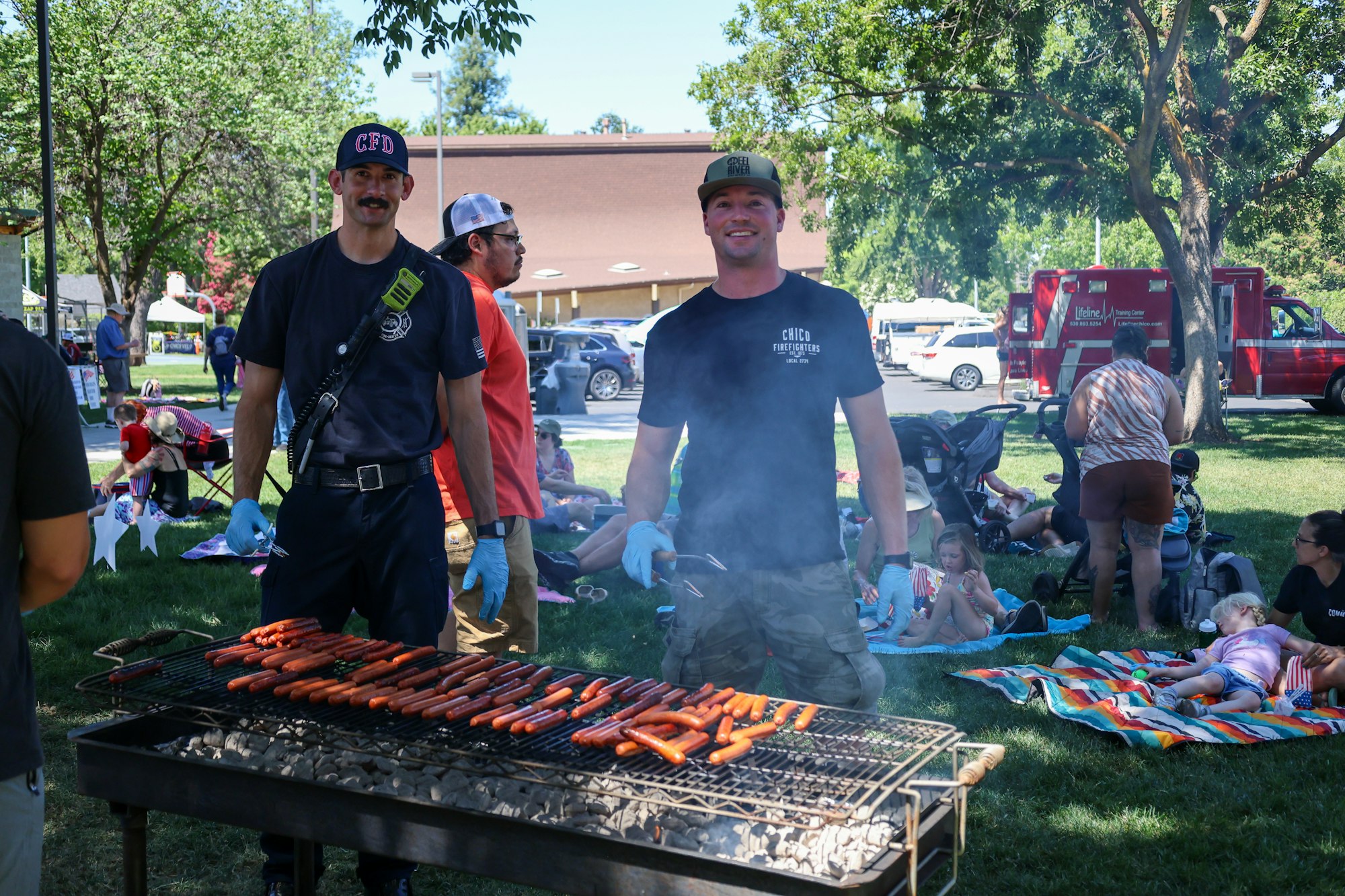 Two people grilling hot dogs at an outdoor gathering with others in the background on a sunny day.