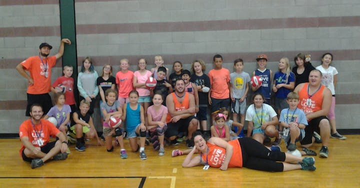 A large group of kids and adults poses together in a gym, some holding volleyballs, all wearing casual sports attire.