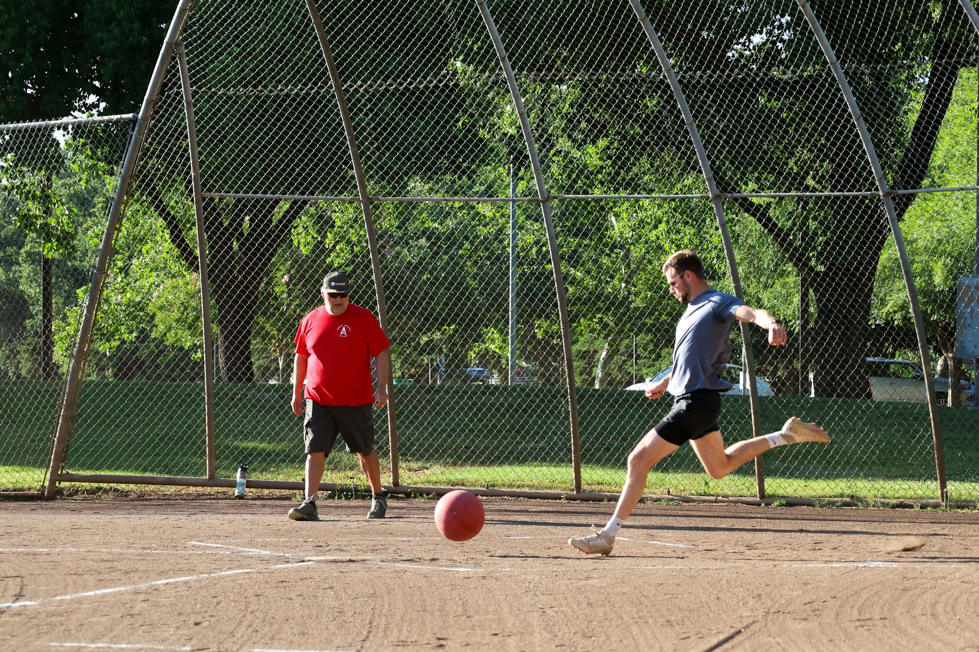 A man kicks a red ball on a dirt field while another person watches behind a fence.