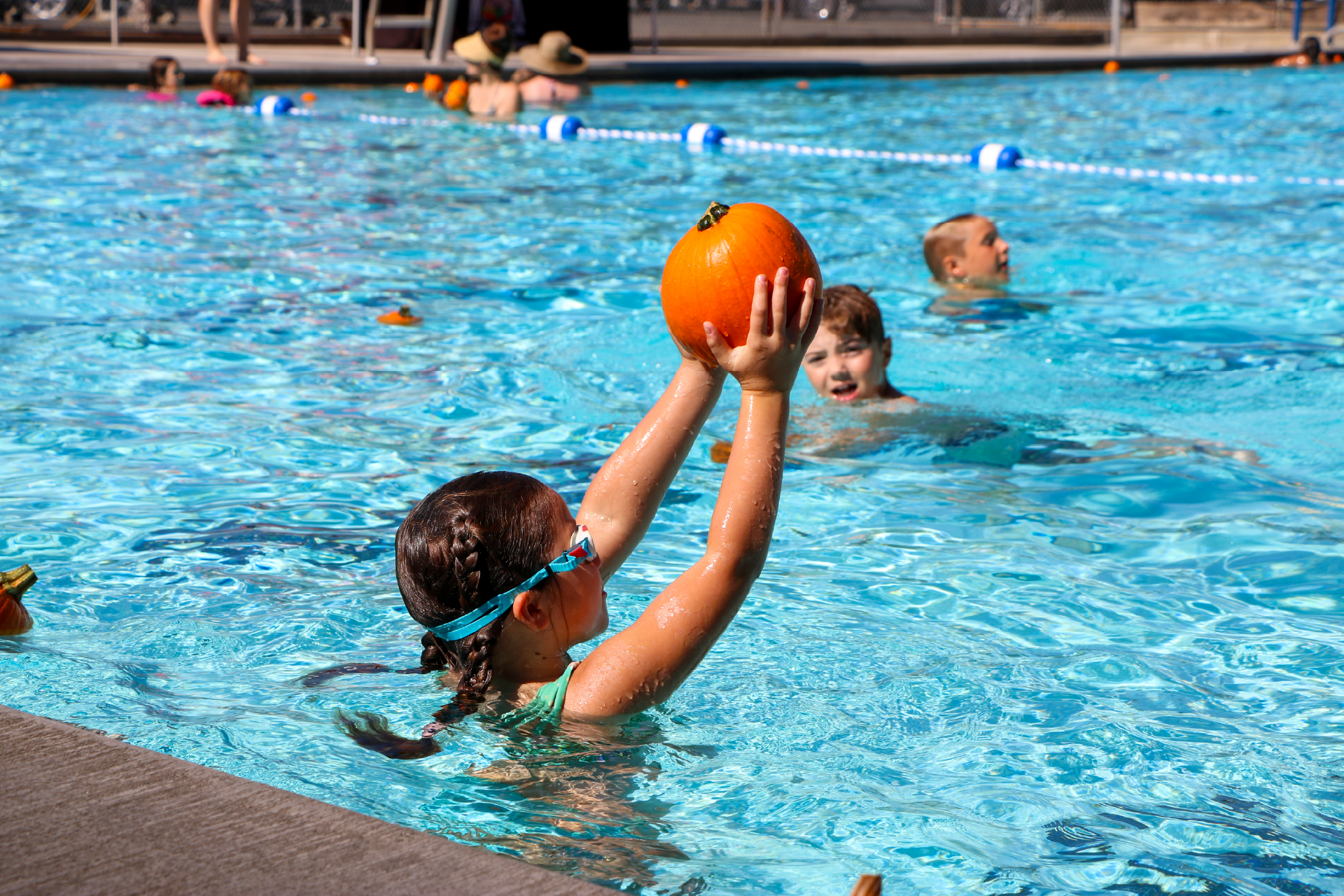 A child holds a pumpkin in a swimming pool with others around.