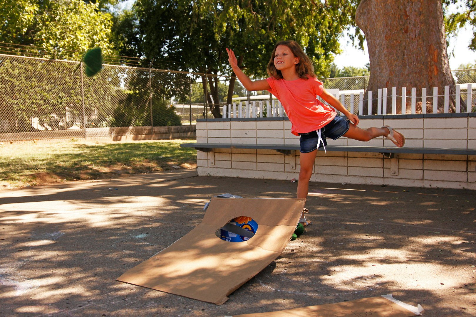 A girl in a bright shirt is playfully tossing a ball towards a cardboard target in an outdoor setting.