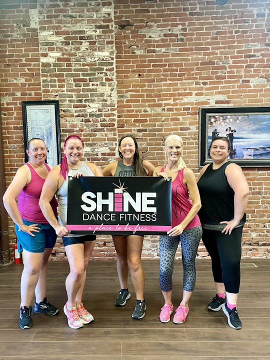 Five women posing with a "SHINE Dance Fitness" sign in a studio with brick walls.