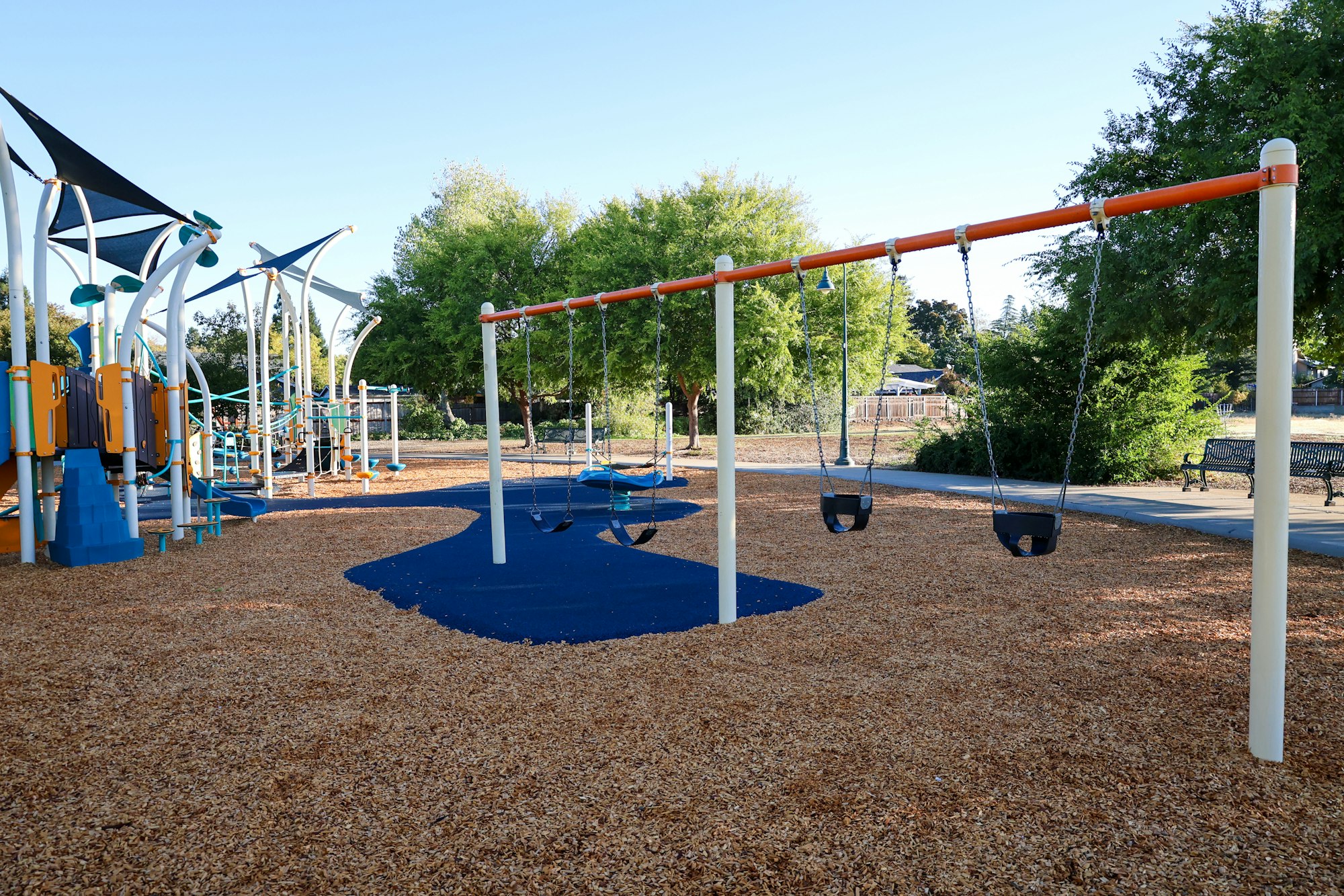 Playground with swings, slides, climbing structures, wood chip ground, and trees in the background.