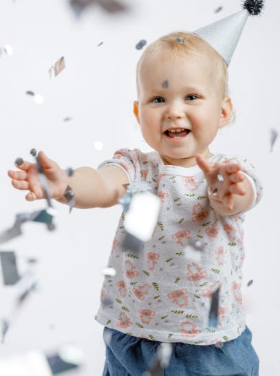 A child wearing a party hat, surrounded by falling confetti, smiling and reaching forward.