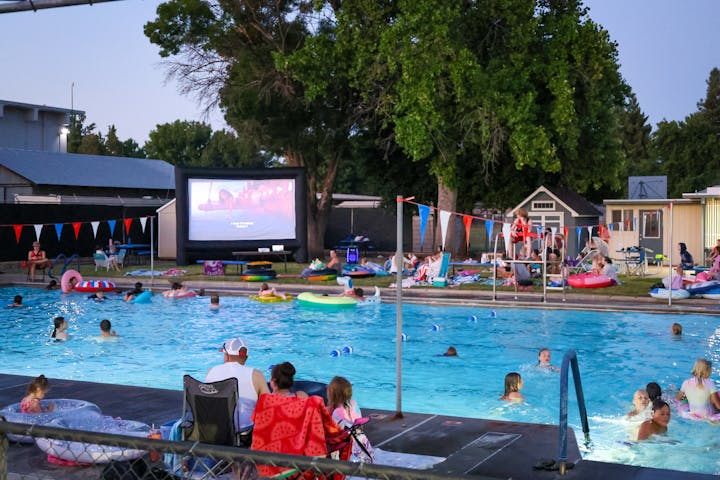 A community pool hosts a movie night with people swimming and relaxing on floats, enjoying the outdoor atmosphere.