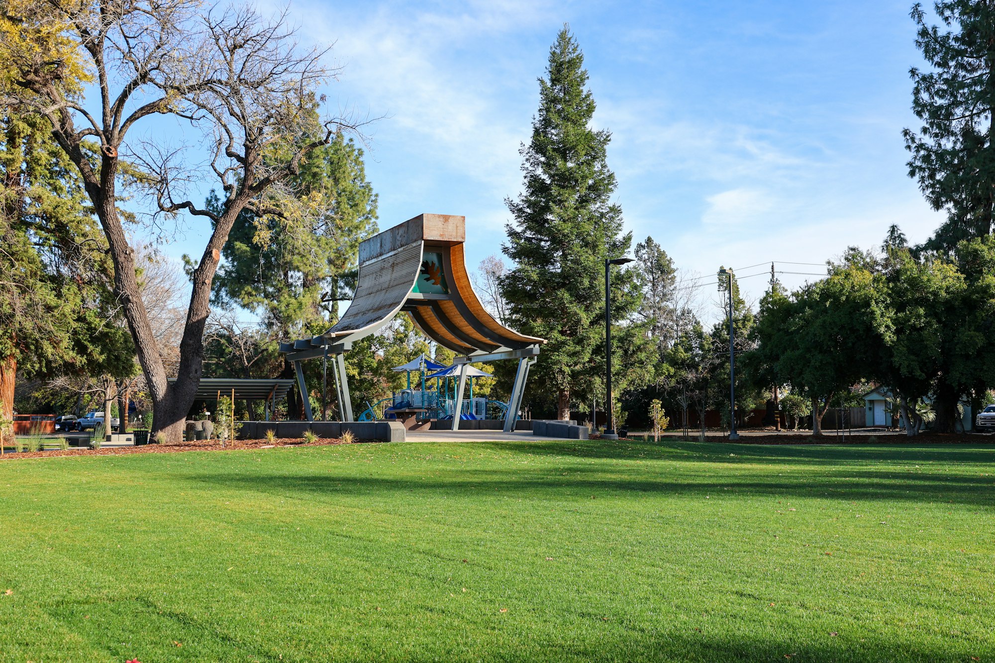 A park with a modern canopy structure, playground, trees, and green grass.