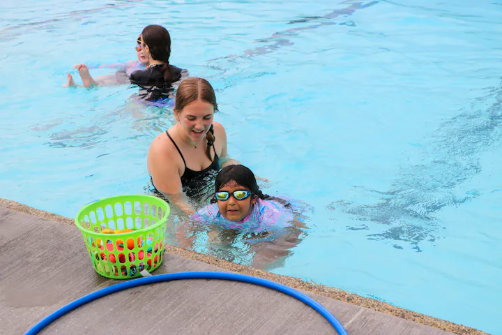 People swimming in a pool, with a basket of pool toys and a blue hose on the deck.