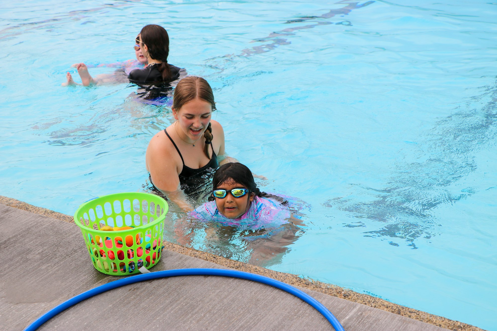 People swimming in a pool, with a basket of pool toys and a blue hose on the deck.