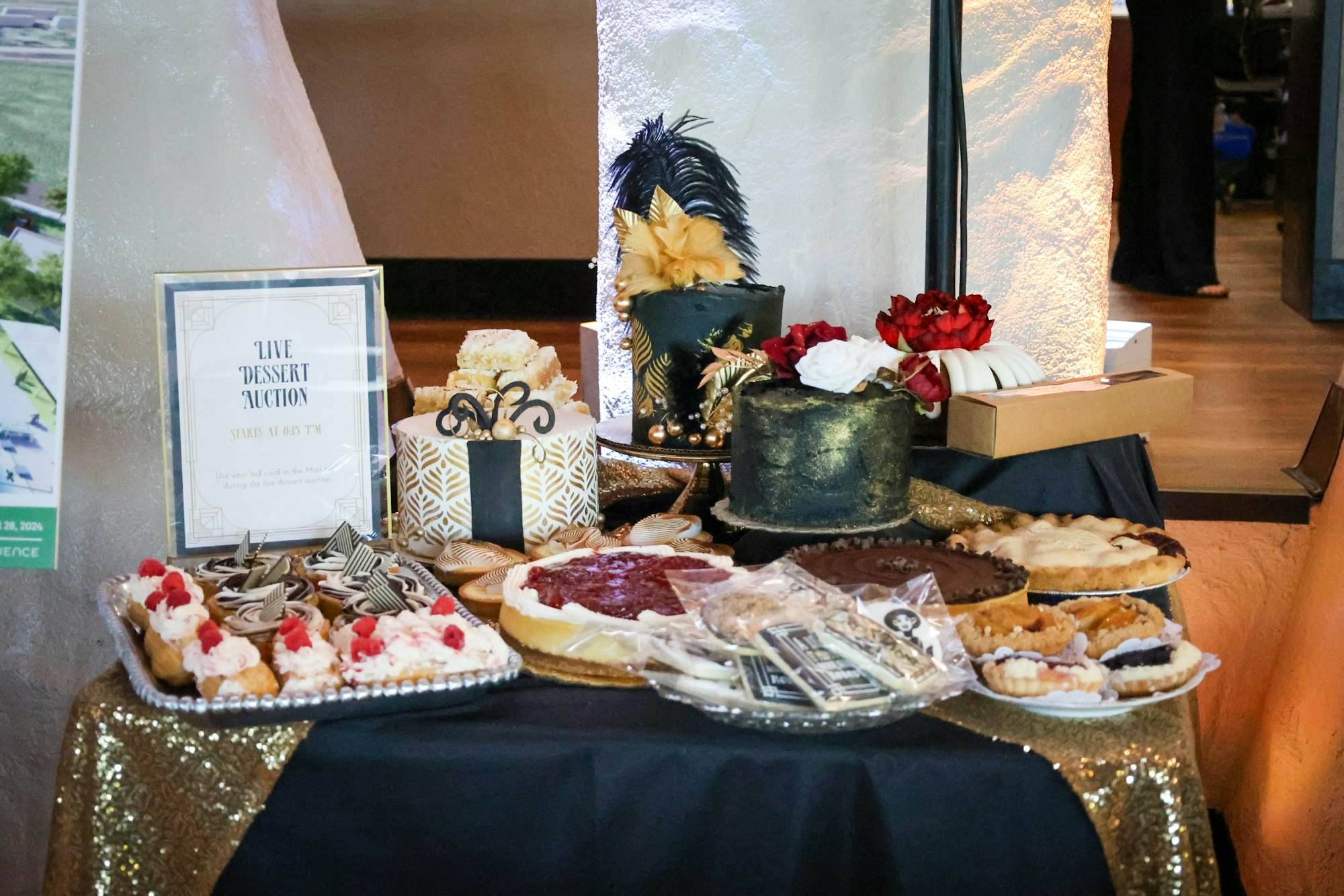A table with assorted desserts, decorated cakes, pies, and a sign for a "Live Dessert Auction" on a sparkling tablecloth.