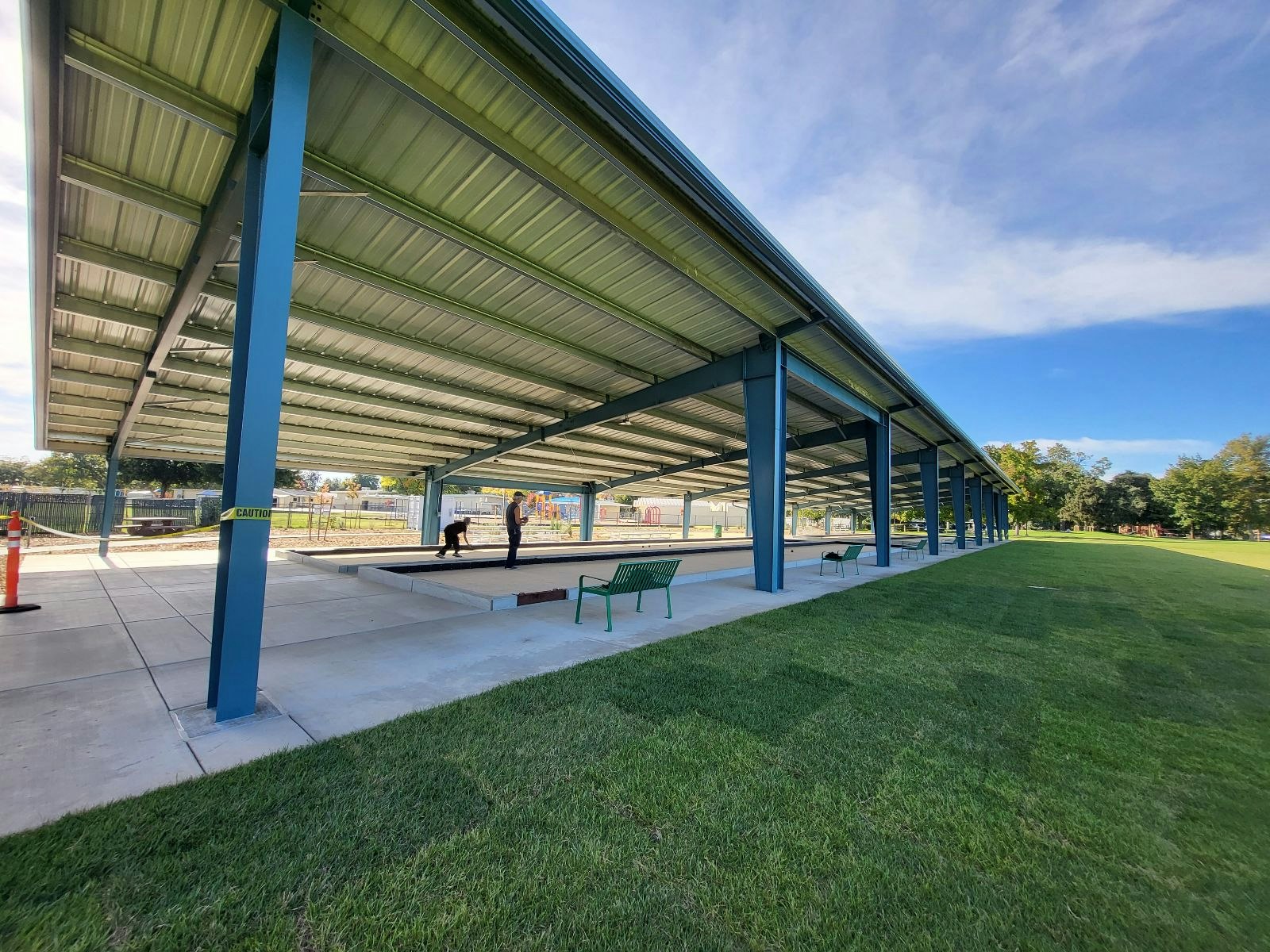 Open-air covered pavilion with benches, green grass, blue sky, and a person at a distance.