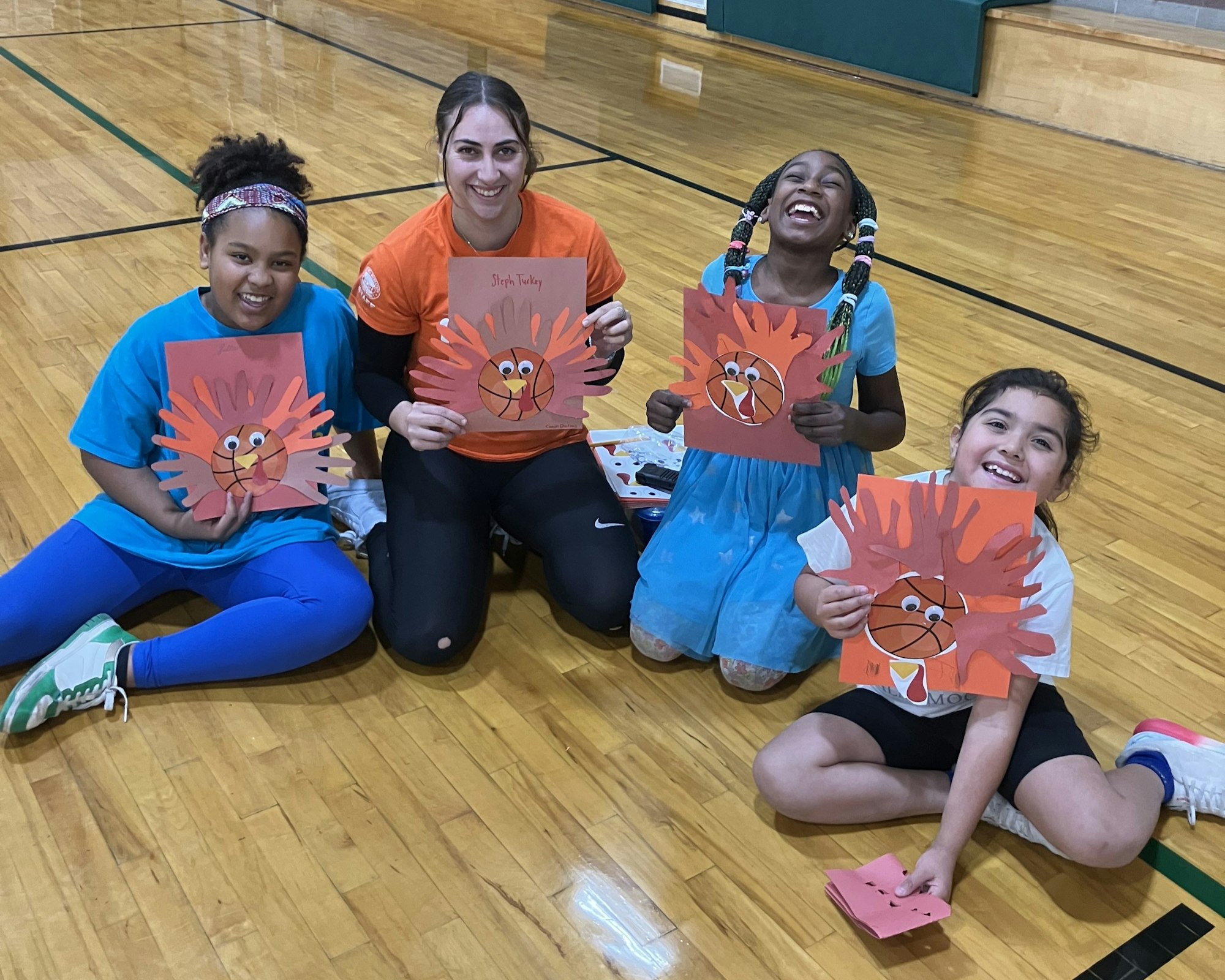 A group of children and an adult smile while holding colorful turkey crafts made from handprints in a gym.